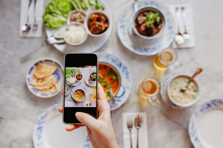 Hand of anonymous female using smartphone to take photos of delicious dishes on table