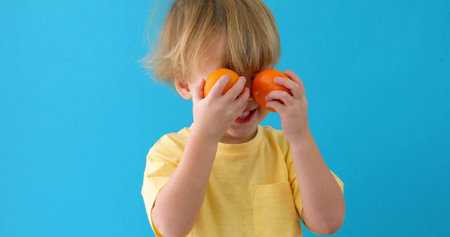 Littele boy plays with mandarins. Kid holds tangerines like eyeglasses blue background