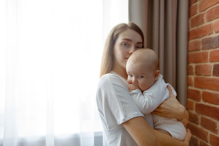 Happy young brunette mother hugging child sucking hand and looking at camera standing near window at homeの写真素材