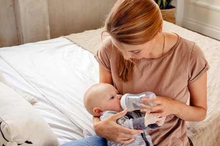 Baby boy drinking milk from bottle holding by mother at homeの写真素材