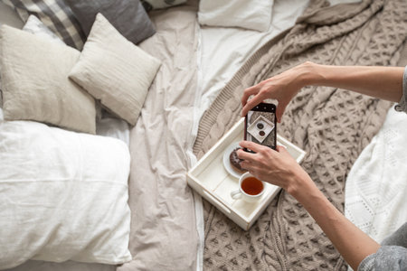 From above shot of woman sitting on bed and taking picture of tea and doughnut on tray with smartphoneの写真素材