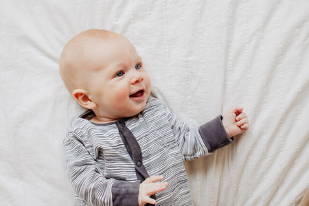 From above adorable baby smiling and looking away while lying on soft bed at home