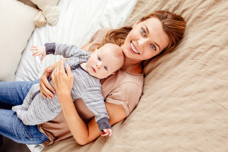 Beautiful young woman with cute baby smiling and looking at camera while lying on comfortable bed at homeの写真素材