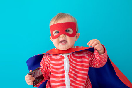 Back view of cute baby in superhero mask and cape looking at camera blue backgroundの写真素材
