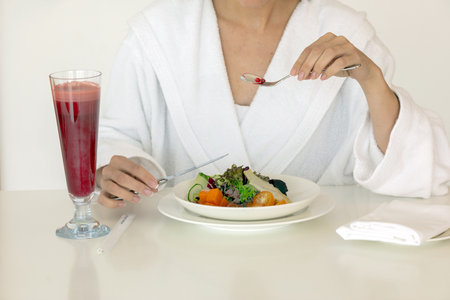 Fresh tasty vegetable salad berry drink and woman in white fluffy robe eating with knife and fork at tableの写真素材