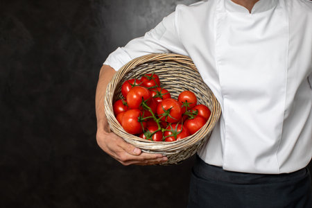 Crop cook in uniform holding basket with fresh ripe tomatoes on black backgroundの写真素材