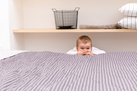 High angle of funny little boy peeking out over opposite side of bed and covering mouth with hands...