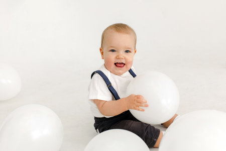 Little vale toddler in white polo and denim jumpsuit laughing while playing with white balloons and...
