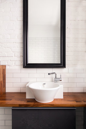 Big mirror with dark frame hanged on white painted and tiled brick wall over wooden counter around ceramic sink in light washroom with interior design in loft styleの写真素材