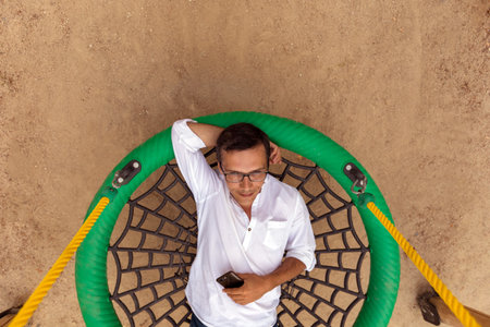 From above of male businessman with smartphone relaxing on round swing in park during working day
