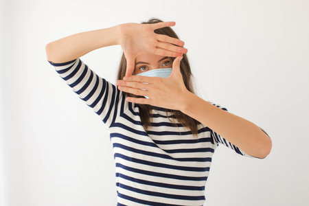 Serious young female in casual striped shirt and protective mask for coronavirus prevention looking at camera through framing hands against white backgroundの写真素材