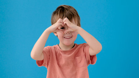 Cute little kid in orange t-shirt makes heart shape with fingers posing for camera on bright...