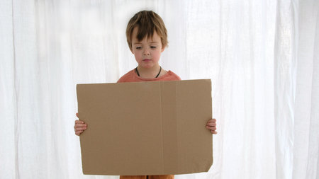 Upset little boy in orange holds large cardboard sheet with space for template standing near white curtain in light roomの写真素材