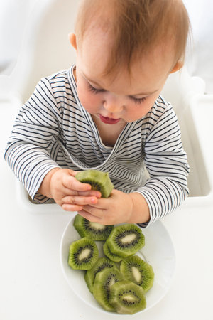 Cute baby girl are sitting whith kiwi white background interior. Funny child explores the fruitの写真素材
