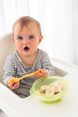 Cute baby girl are sitting whith sliced banana white background interior. Funny child explores the fruitの写真素材