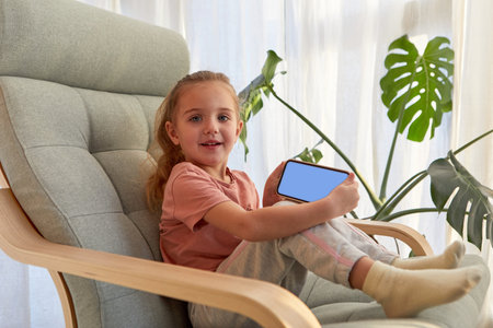 Cute child showing mobile phone while sitting on armchairの写真素材