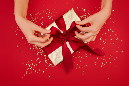 Female hands tying a bow on gift box with a red ribbon on wine background. Concept of a gift for the...