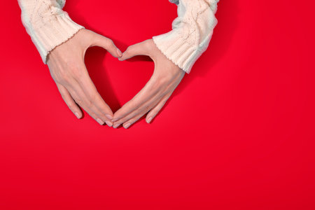 Top view of anonymous person in sweater showing heart shaped gesture with hands against red backgroundの写真素材