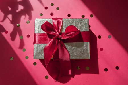 Gift golden wrapped box with a bow on a red background with confetti in hard light with shadows from a home plant. Top view present box package over flat lay backgroundの写真素材