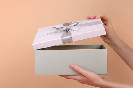 Side view of hands opening a box with a gift bow on a peach background