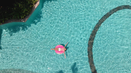 Young female freelancer sitting with a tablet in an inflatable circle in the pool. Aerial view