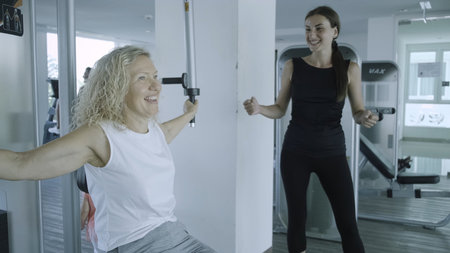 Senior woman is engaged on a simulator in the gym with a personal trainer. daughter helps mom in the gymの写真素材