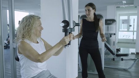 Senior woman is engaged on a simulator in the gym with a personal trainer. daughter helps mom in the gymの写真素材