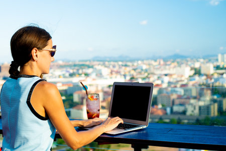 The girl works on a computer in a cafe on the roof. Female writing on a laptop in a rooftop cafe, sitting at a wooden table with a panoramic view of the city.の写真素材