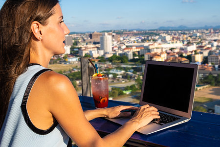 The girl works on a computer in a cafe on the roof. Female writing on a laptop in a rooftop cafe, sitting at a wooden table with a panoramic view of the city.の写真素材