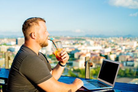 man drinking a cocktail and working on a computer in a cafe with a beautiful viewの写真素材