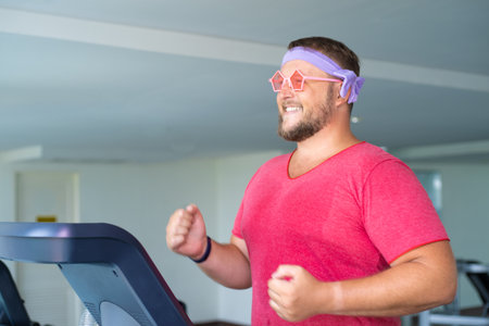 Playful fat man in a pink T-shirt and pink glasses is engaged in fitness in the gym.の写真素材