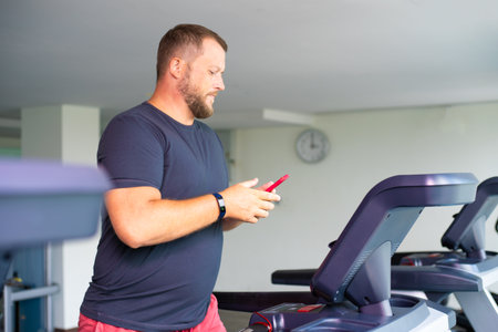 chubby man walking on running track, warming up on gym treadmill. man with phoneの写真素材