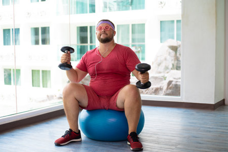Playful fat man in a pink T-shirt and pink glasses is engaged in fitness with dumbbells and a fit ball in the gym. man drinking water in the gym. man listening to music on headphonesの写真素材