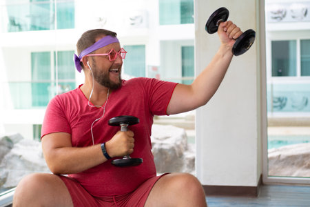 Playful fat man in a pink T-shirt and pink glasses is engaged in fitness with dumbbells and a fit ball in the gym. man drinking water in the gym. man listening to music on headphonesの写真素材