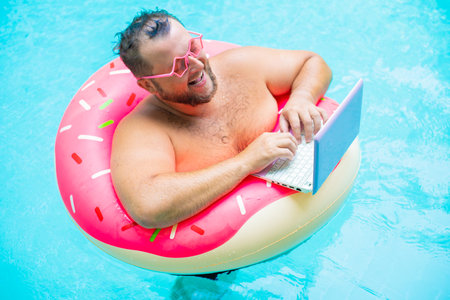 smiling Funny fat male in pink glasses on an inflatable circle in the pool works on a laptop portraying a girl.の写真素材
