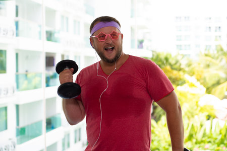 Playful fat man in a pink T-shirt and pink glasses is engaged in fitness in the gym. man drinking water in the gymの写真素材