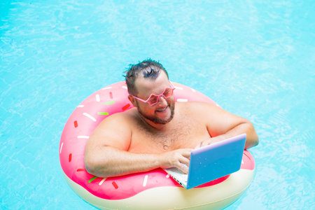 enjoying Funny fat male in pink glasses on an inflatable circle in the pool works on a laptop portraying a girl.の写真素材