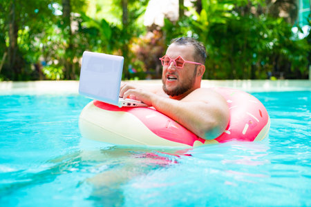 surprised Funny fat male in pink glasses on an inflatable circle in the pool works on a laptop portraying a girl.の写真素材