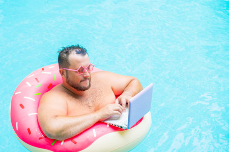serious Funny fat male in pink glasses on an inflatable circle in the pool works on a laptop portraying a girl.の写真素材