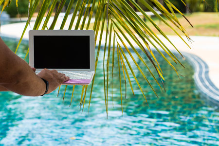 A man is typing on his laptop while sitting by the pool. Mens hands on the keyboard in the poolの写真素材