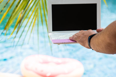 A man is typing on his laptop while sitting by the pool. Mens hands on the keyboard in the poolの写真素材