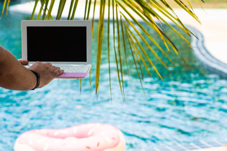 A man is typing on his laptop while sitting by the pool. Mens hands on the keyboard in the poolの写真素材
