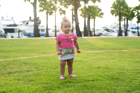 A little girl of 1 year old running on a green meadow in a park among palm trees and cutting a red ribbonの写真素材