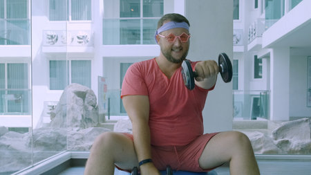Playful fat man in a pink T-shirt and pink glasses is engaged in fitness with dumbbells and a fit ball in the gymの写真素材