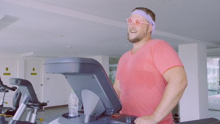 Playful fat man in a pink T-shirt and pink glasses is engaged in fitness in the gym.