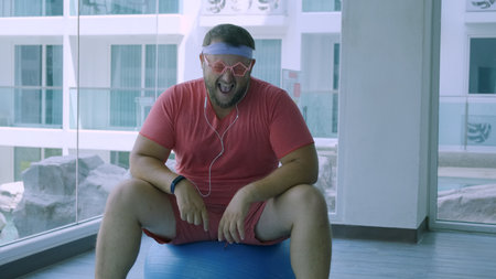 Playful fat man in a pink T-shirt and pink glasses is engaged in fitness with dumbbells and a fit ball in the gym. man listening to music on headphones.の写真素材