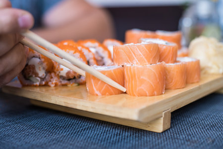 Hand with wooden chopsticks takes rolls that stand on a wooden stand and blue litter, blurred background, close up.の写真素材