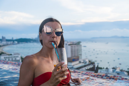 young attractive girl with glasses stands in a rooftop pool of a skyscraper with a cocktail and...