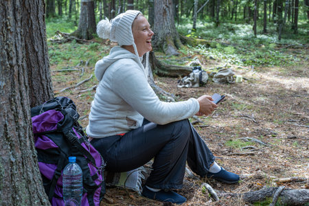 A pretty girl in light clothes, a European-looking ecotourist, sits near a tree in the forest and looks at the phone.の写真素材