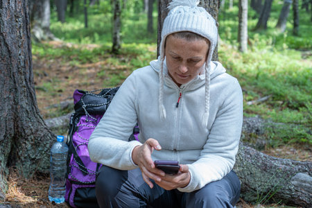 A pretty girl in light clothes, a European-looking ecotourist, sits near a tree in the forest and looks at the phone.の写真素材
